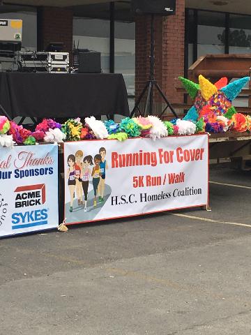Banner reading "Running for Cover 5K Run / Walk, HSC Homeless Coalition" hanging on the front of the stage.