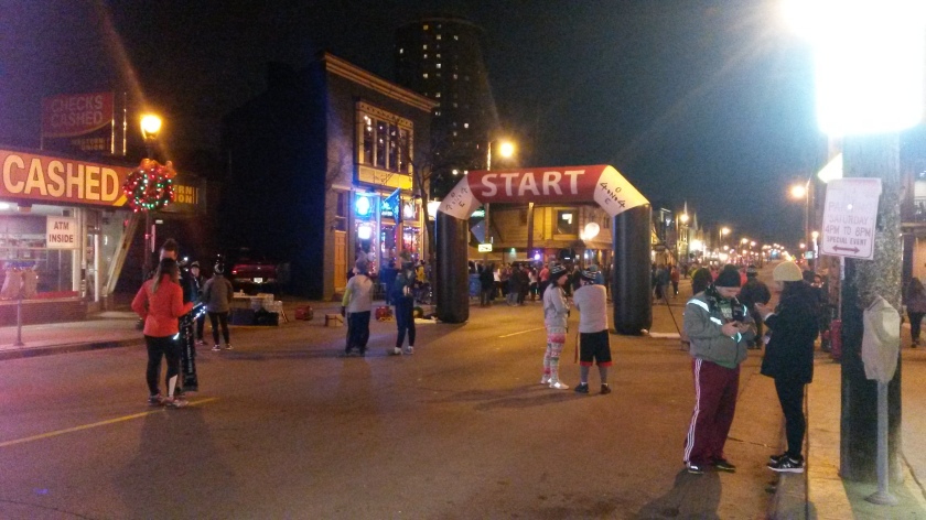 Nighttime; a sparse crowd of runners by an inflatable "Start" arch in the middle of a city street with assorted Christmas decorations.