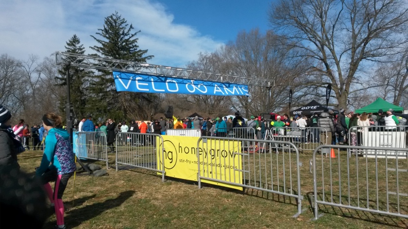 Runners milling around outside the barricades of the race starting area. A blue banner reading "Velo Amis" is stretched out above the starting line.
